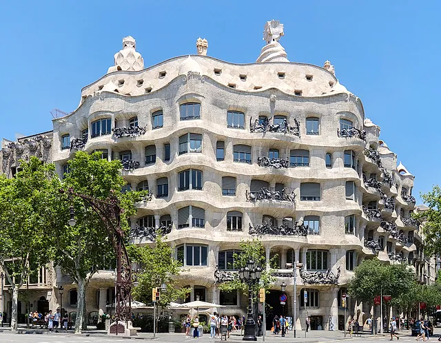 The undulating stone facade of Casa Milà seen from Passeig de Gràcia on a clear day, with its wrought-iron balconies and the plane trees along the pavement