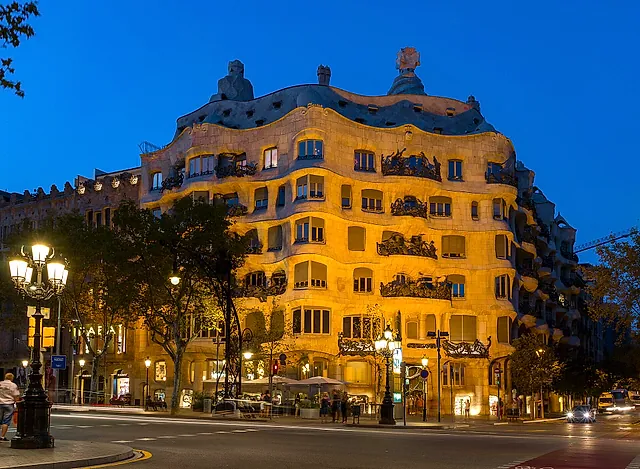 Casa Milà lit by warm light at dusk, with the wave-shaped facade glowing above the street and its lamp posts